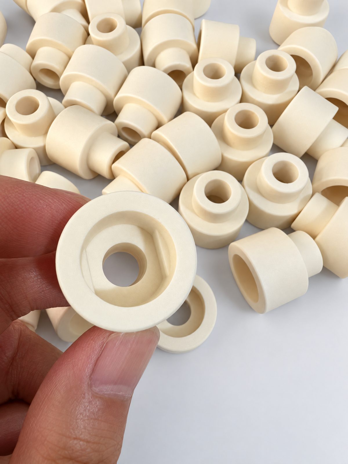 A pile of cream stepped ceramic bushings with through bores; a hand holds one to show the counterbore in its larger end.
