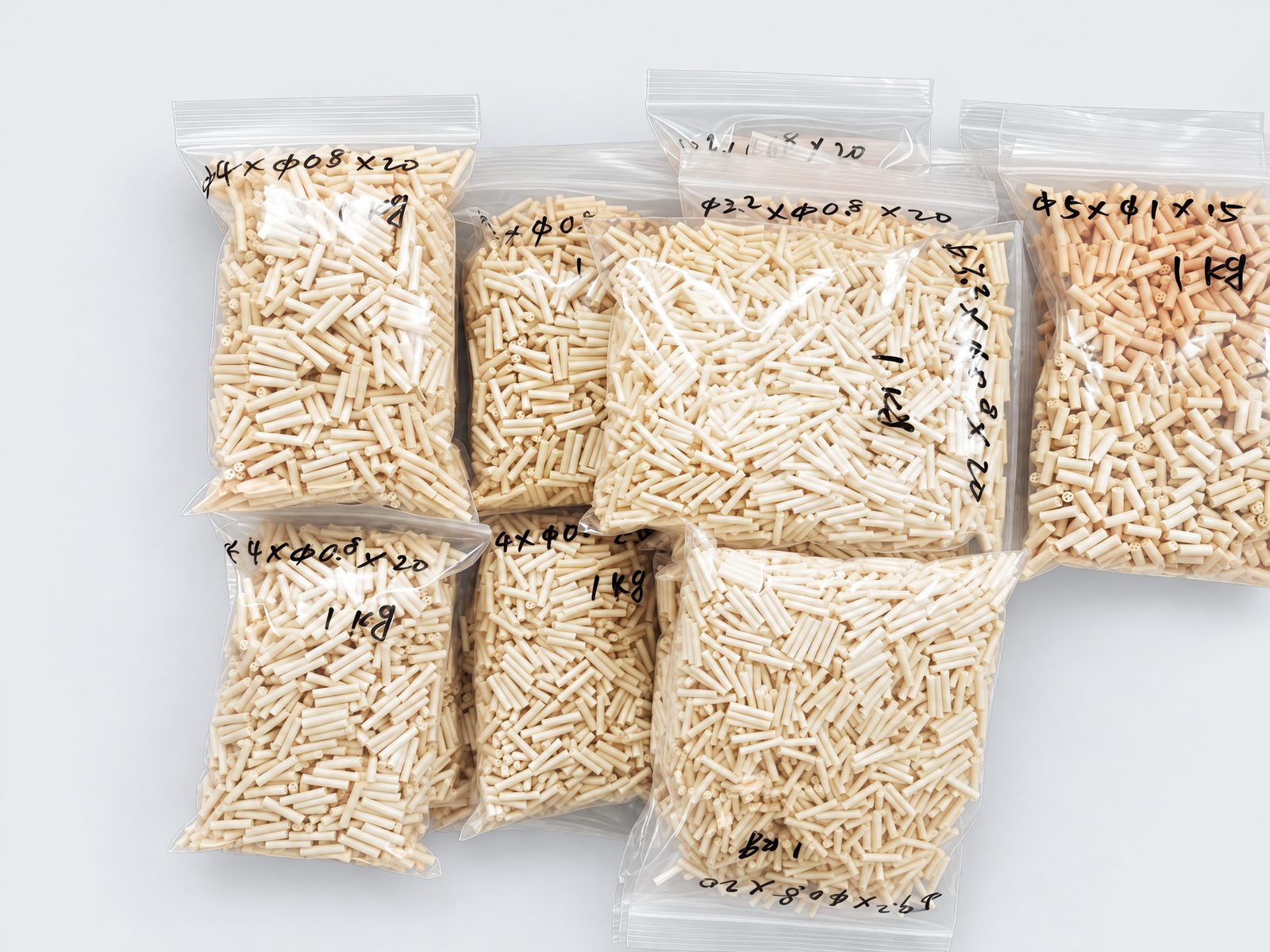 Zip-lock bags of short cream multi-bore beads, each marked by hand with its size and 1 kg, on a plain grey background.