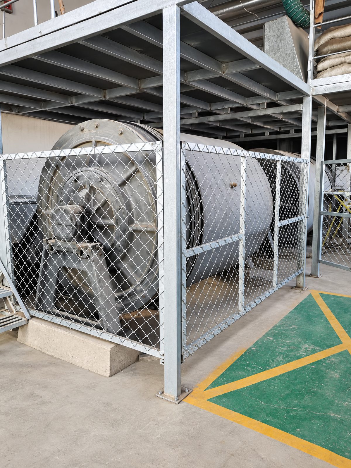 Two large grey drum ball mills inside a galvanised steel frame with diamond-mesh safety panels, under a steel mezzanine.